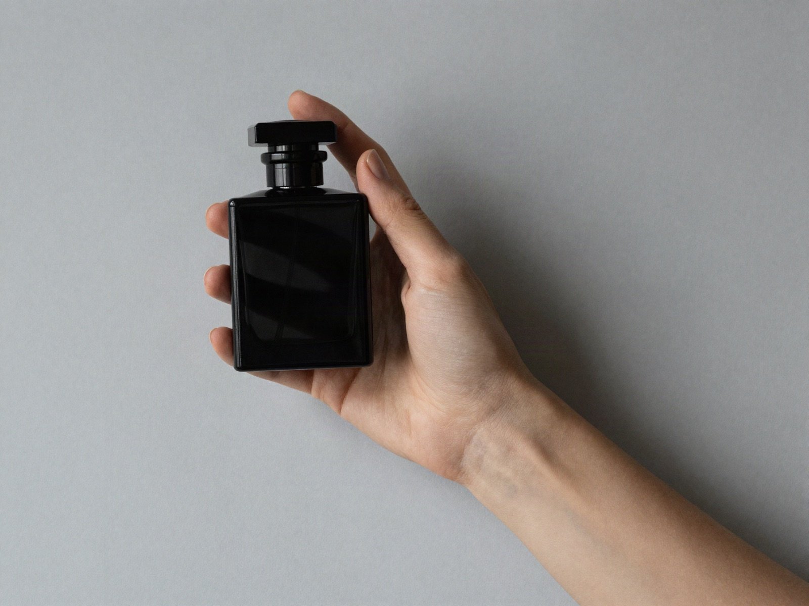 يد تحمل زجاجة عطر سوداء مربعة أمام جدار رمادي